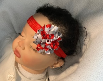 May include: A red headband with a gray, white, and red bow. The bow is made of ribbon and is tied in a knot. The headband is worn by a baby.