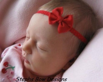 Può includere: Un fiocco in tessuto rosso attaccato a una sottile fascia elastica rossa testurizzata. La fascia è indossata su una testa e l'indumento visibile è una maglia rosa chiaro con un motivo a ciliegie rosse e il testo Stephs Bow Designs.