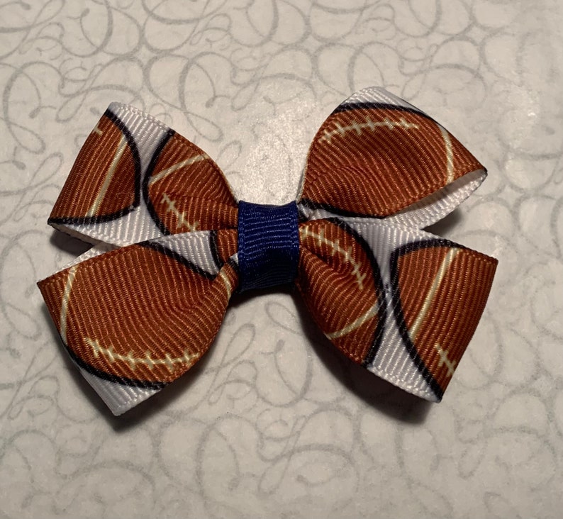 Puede incluir: Un lazo para el cabello hecho a mano con un dise&ntilde;o de f&uacute;tbol americano. El lazo est&aacute; hecho de cinta marr&oacute;n y blanca con un patr&oacute;n de f&uacute;tbol americano y un nudo central azul marino. Un accesorio deportivo.