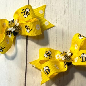 May include: Two bright yellow hair bows made of ribbed fabric, each featuring a repeating pattern of small black and yellow bees and white flowers. A three-dimensional bee charm is attached to the center of each bow.