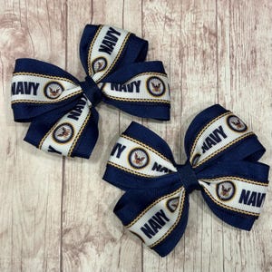 May include: Two hair bows made of navy blue and white grosgrain ribbon. The white center stripe features the repeating text NAVY in black capital letters and a circular emblem with a golden eagle. The edges are trimmed with thin gold lines.