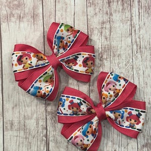 May include: Two pink hair bows with a white ribbon center featuring colorful cartoon characters. The bows are made of grosgrain ribbon and are arranged on a light brown wooden surface.