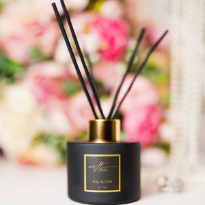 May include: A black diffuser bottle with a gold collar and black reeds. The label reads "Full Bloom" and "4oz (114g)". The background is a soft focus of pink and white flowers, suggesting a floral fragrance.