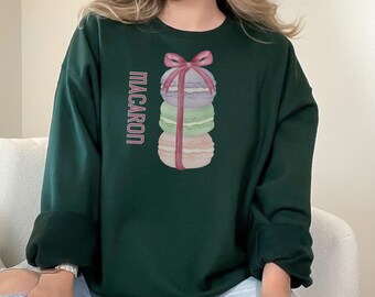 May include: A dark green sweatshirt with a stack of pastel macarons tied with a red ribbon. The text "MACARON" is printed on the left side of the sweatshirt.