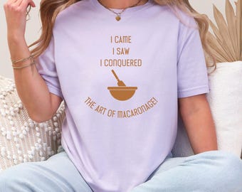 Puede incluir: Camiseta lila claro con el texto "I CAME I SAW I CONQUERED" sobre un gráfico de un cuenco y un batidor. Debajo del gráfico está el texto "THE ART OF MACARONAGE!"