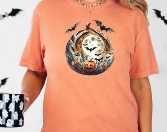 May include: Orange t-shirt featuring a Halloween-themed graphic. The design includes a full moon, bats, and pumpkins. The shirt is a short-sleeved, crew neck style. The model is wearing a necklace and a black and white checkered skirt.
