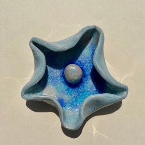 May include: A blue and white ceramic dish shaped like a star with a small white and iridescent round knob in the center.