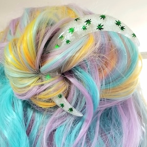 May include: A white hair clip with a green glitter design of marijuana leaves. The clip is shaped like a crescent moon.