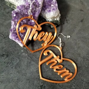 May include: A pair of copper-colored heart-shaped earrings with the words "They" and "Them" cut out in the center. The earrings are hanging from silver hooks.