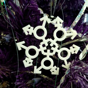 May include: White snowflake ornament with a transgender symbol design on a purple Christmas tree.