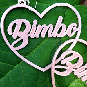 K&ouml;nnte beinhalten: Ein Paar rosa herzf&ouml;rmige Ohrringe mit dem Wort "Bimbo" in kursivschrift.