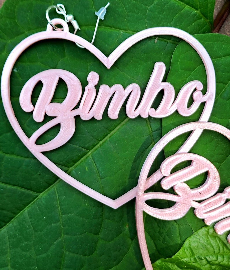 K&ouml;nnte beinhalten: Ein Paar rosa herzf&ouml;rmige Ohrringe mit dem Wort "Bimbo" in kursivschrift.