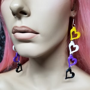 May include: A pair of dangle earrings with four layered hearts. The hearts are black, purple, white, and yellow. The earrings are made of plastic and have silver hooks.