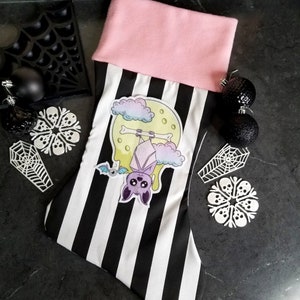 May include: A black and white striped Christmas stocking with a pink cuff. The stocking features an applique of a purple bat hanging from a bone against a yellow moon with clouds.