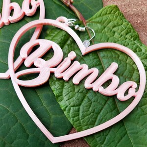 K&ouml;nnte beinhalten: Rosa herzf&ouml;rmige Ohrringe mit dem Wort "Bimbo" in kursivschrift.
