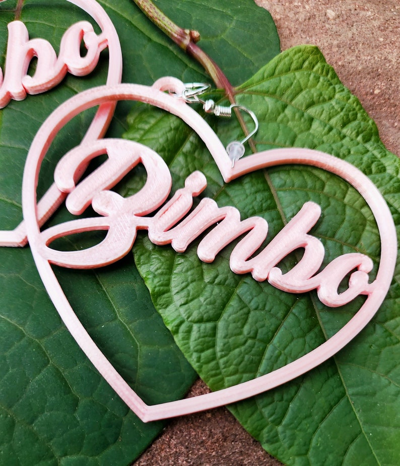 K&ouml;nnte beinhalten: Rosa herzf&ouml;rmige Ohrringe mit dem Wort "Bimbo" in kursivschrift.