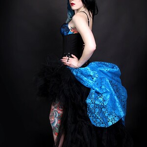 May include: A woman with blue and black hair wearing a black corset and a blue and black tulle skirt. She has a tattoo on her leg.
