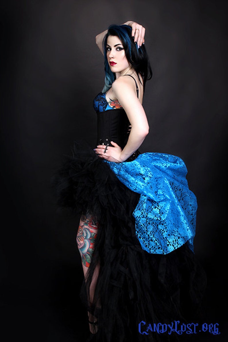 May include: A woman with blue and black hair wearing a black corset and a blue and black tulle skirt. She has a tattoo on her leg.