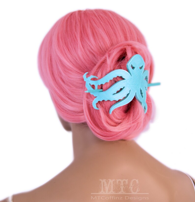 May include: A light blue octopus hair clip with a textured surface. The clip is in the hair of a person with pink hair.