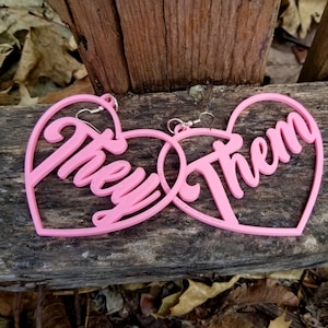 May include: Pink heart-shaped earrings with the words "They" and "Them" written in a stylized font.