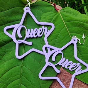 May include: A pair of lavender star-shaped earrings with the word "Queer" written in a cursive font on each earring.