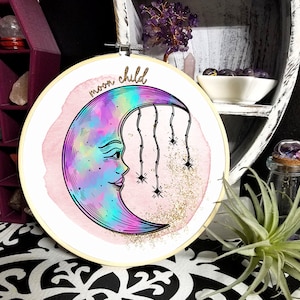 May include: A pink and white embroidery hoop with a crescent moon design. The moon is iridescent and has a face. The text "moon child" is written above the moon.
