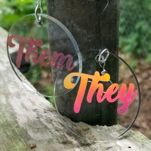 May include: A pair of clear acrylic earrings with a gradient pink and orange design. The earrings are round and feature the words "Them" and "They" in a stylized font.
