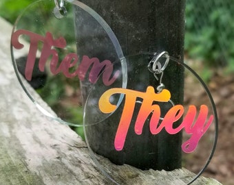 May include: A pair of clear acrylic earrings with a gradient pink and orange design. The earrings are round and feature the words "Them" and "They" in a stylized font.