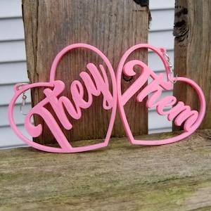 May include: A pair of pink heart-shaped earrings with the words "They" and "Them" written in a cursive font.