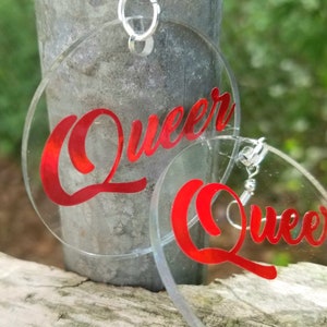 May include: A pair of clear acrylic earrings with red vinyl lettering that says "Queer".