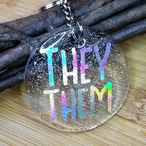 May include: A round, clear keychain with iridescent glitter embedded in the resin. The keychain features the words "THEY THEM" in rainbow colors.