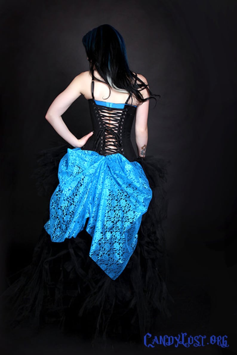 May include: A woman wearing a black corset with a blue lace overlay and a black tulle skirt. The corset has a lace-up back.