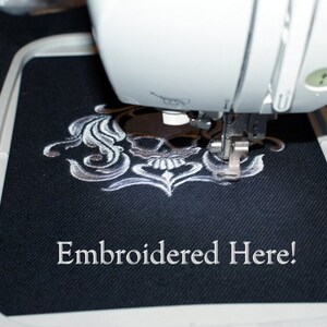 Puede incluir: Primer plano de un dise&ntilde;o de bordado de calavera blanca y flores que se est&aacute; cosiendo en una tela negra con una m&aacute;quina de coser. El texto "Embroidered Here!" es visible debajo del dise&ntilde;o.