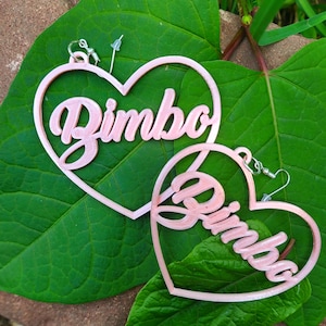 K&ouml;nnte beinhalten: Ein Paar rosa herzf&ouml;rmige Ohrringe mit dem Wort "Bimbo" in Kursivschrift auf jedem Ohrring.