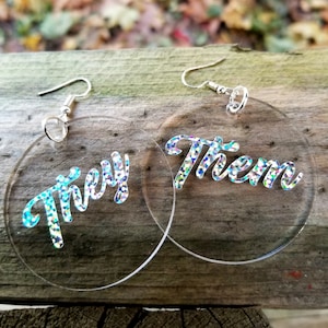 May include: A pair of clear acrylic earrings with silver hooks. The earrings are round and have the words "They" and "Them" written in silver glitter on them.