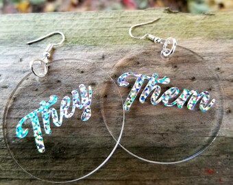 May include: A pair of clear acrylic earrings with silver hooks. The earrings are round and have the words "They" and "Them" written in silver glitter on them.