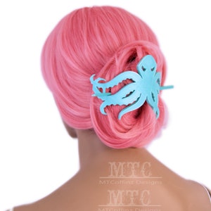 May include: A light blue octopus hair clip with a textured surface. The clip is in the hair of a person with pink hair.