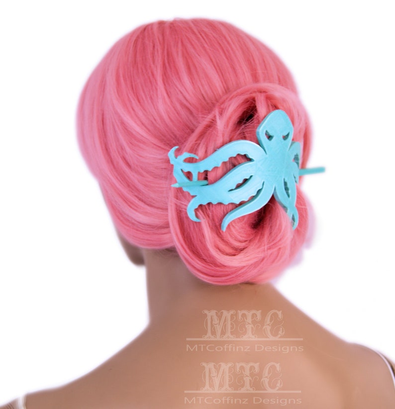 May include: A light blue octopus hair clip with a textured surface. The clip is in the hair of a person with pink hair.