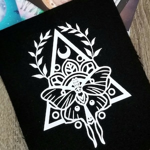 May include: White embroidered patch on black fabric featuring a moth with intricate wings inside a triangle with a crescent moon and floral details.