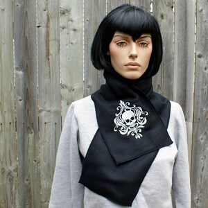 Puede incluir: Bufanda de punto negra con un dise&ntilde;o blanco de calavera y flores. La bufanda est&aacute; doblada y se lleva alrededor del cuello.
