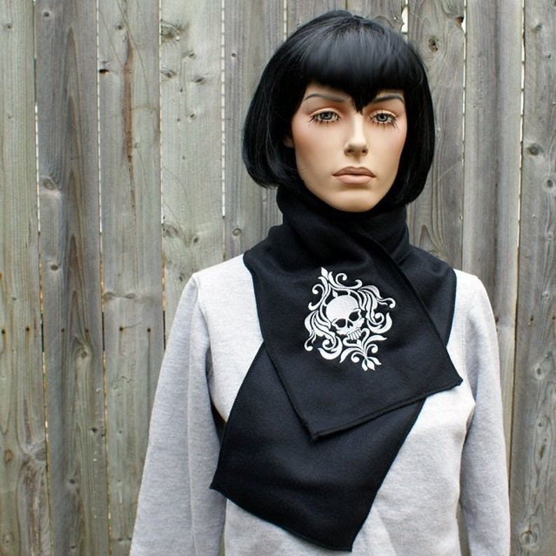 Puede incluir: Bufanda de punto negra con un dise&ntilde;o blanco de calavera y flores. La bufanda est&aacute; doblada y se lleva alrededor del cuello.