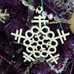 May include: A white snowflake ornament with a unique design hangs from a black string. The ornament is suspended against a backdrop of a purple Christmas tree with decorative beads and lights. The snowflake has a detailed, openwork pattern.