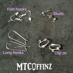 K&ouml;nnte beinhalten: Eine Auswahl an Ohrring-Verschl&uuml;ssen, darunter Fischhaken, Ohrstecker, lange Haken und Clip-Ohrringe. Die Verschl&uuml;sse sind silberfarben und werden auf einer grauen, strukturierten Oberfl&auml;che pr&auml;sentiert.