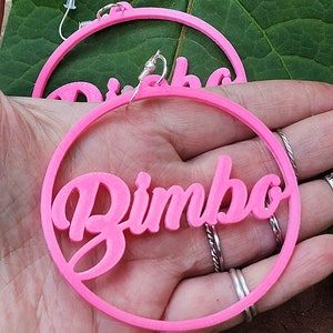 May include: A pair of pink hoop earrings with the word "Bimbo" written in a cursive font.