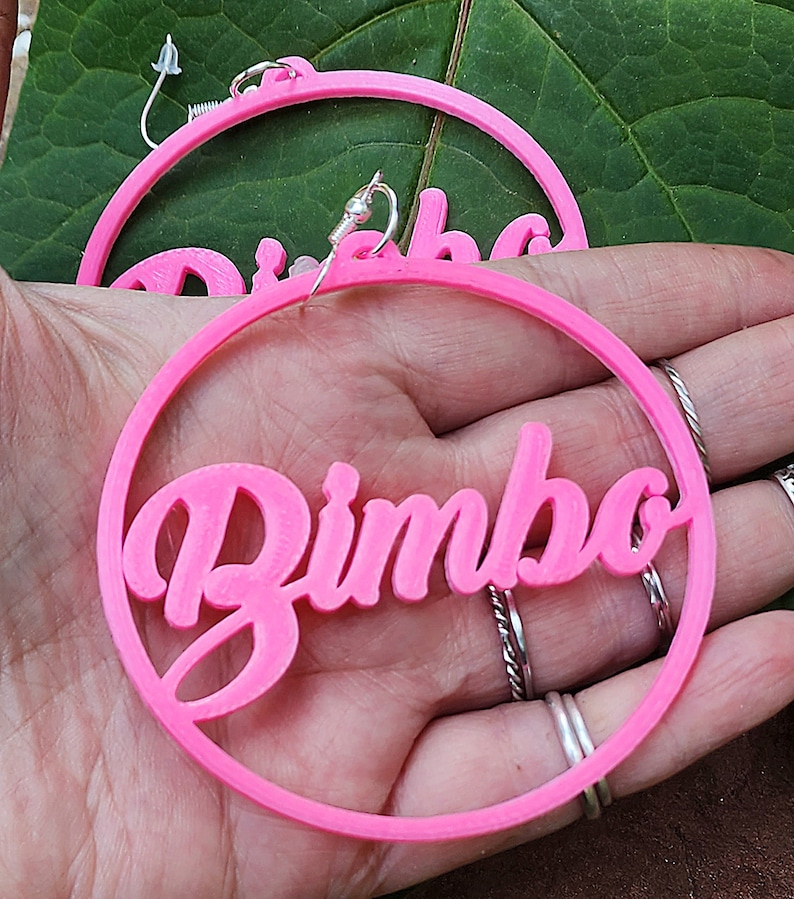May include: A pair of pink hoop earrings with the word "Bimbo" written in a cursive font.