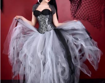 May include: A woman wearing a black and silver corset with a white tulle skirt. The skirt is very full and has a layered look. The woman is standing in front of a red background.