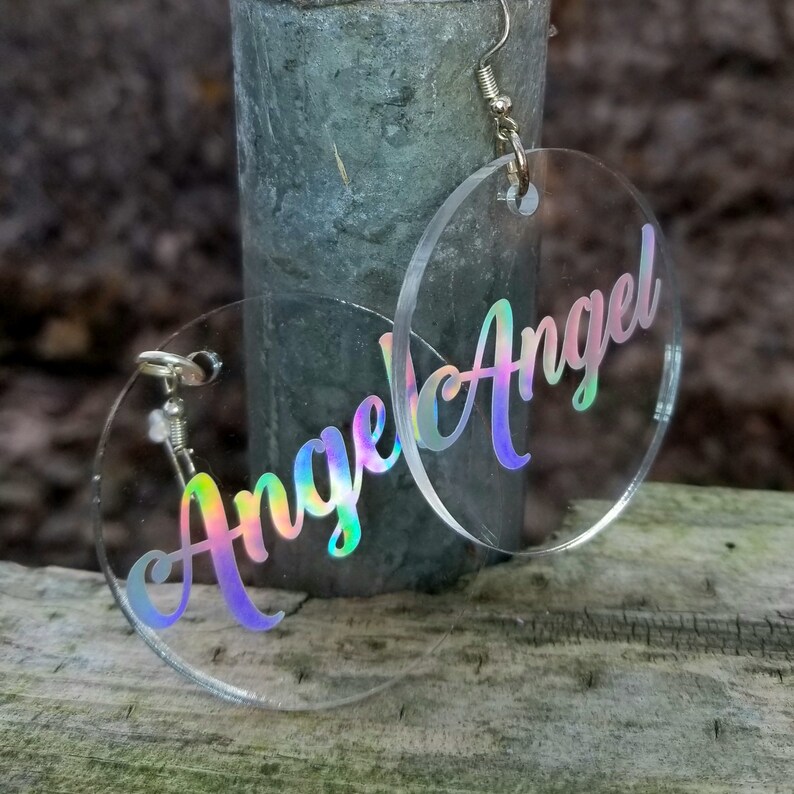 May include: A pair of iridescent clear acrylic earrings with the word "Angel" written in a cursive font.