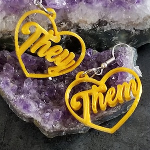May include: A pair of gold heart-shaped earrings with the words "They" and "Them" cut out. The earrings are on a purple background.
