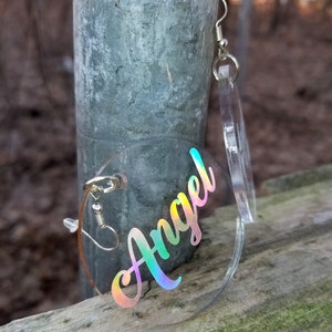May include: A pair of iridescent clear acrylic earrings with the word "Angel" written in a rainbow holographic font.