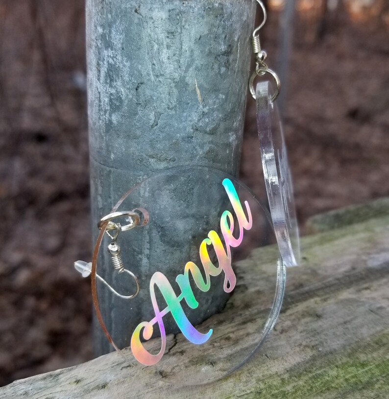 May include: A pair of iridescent clear acrylic earrings with the word "Angel" written in a rainbow holographic font.